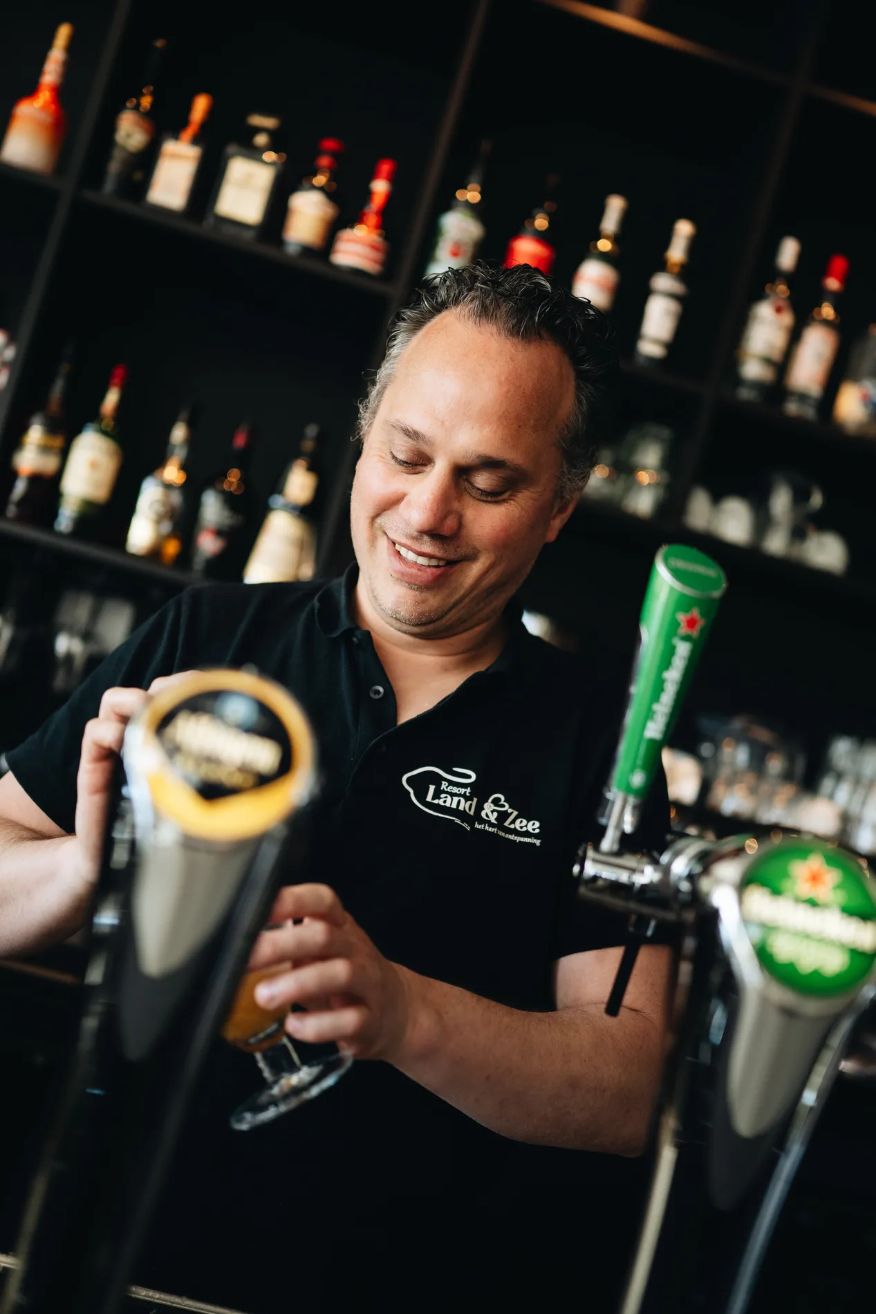 Een glimlachende barman in een zwart shirt met het logo van Resort Land & Zee die een biertje tapt achter de bar, met flessen drank op de achtergrond.